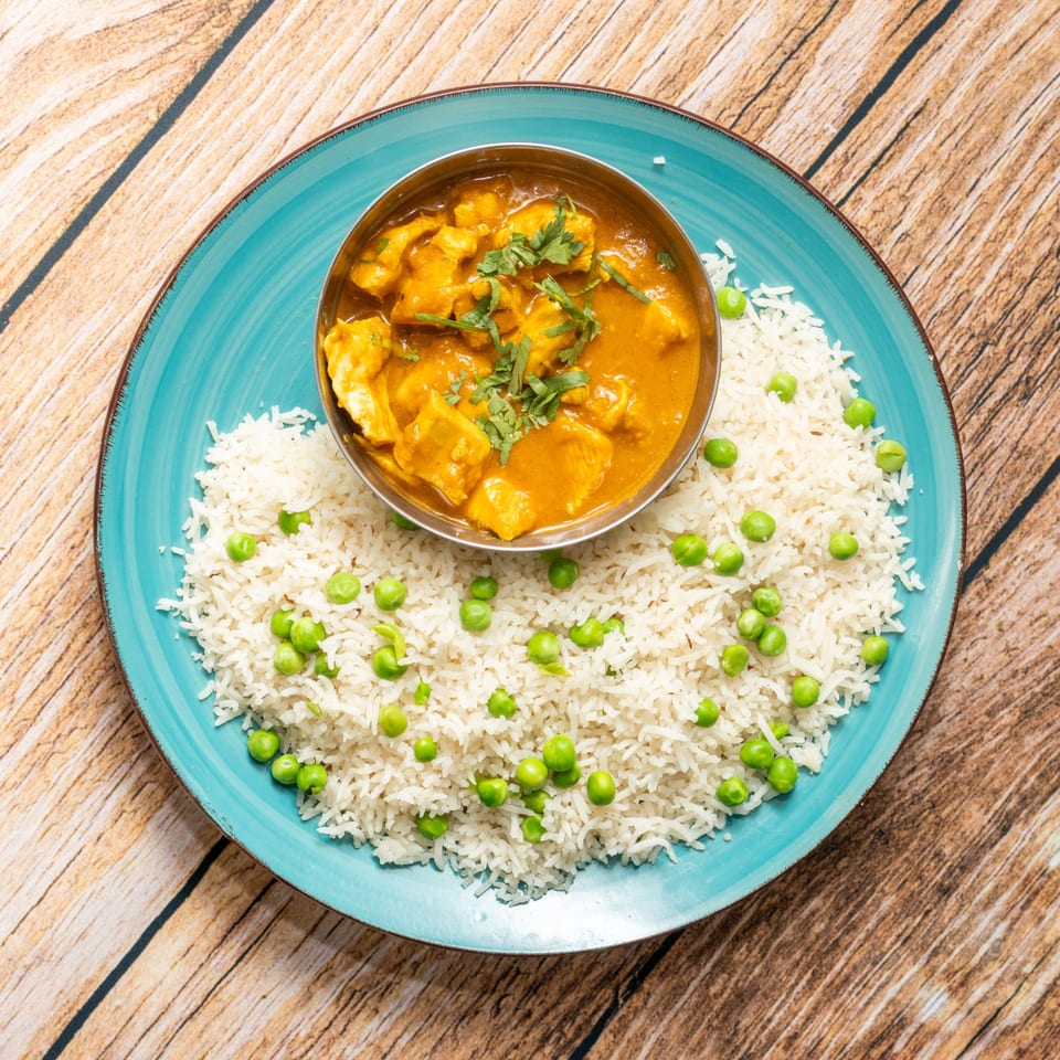 Chicken curry in a metal bowl, surrounded by white rice and green peas on a blue plate.