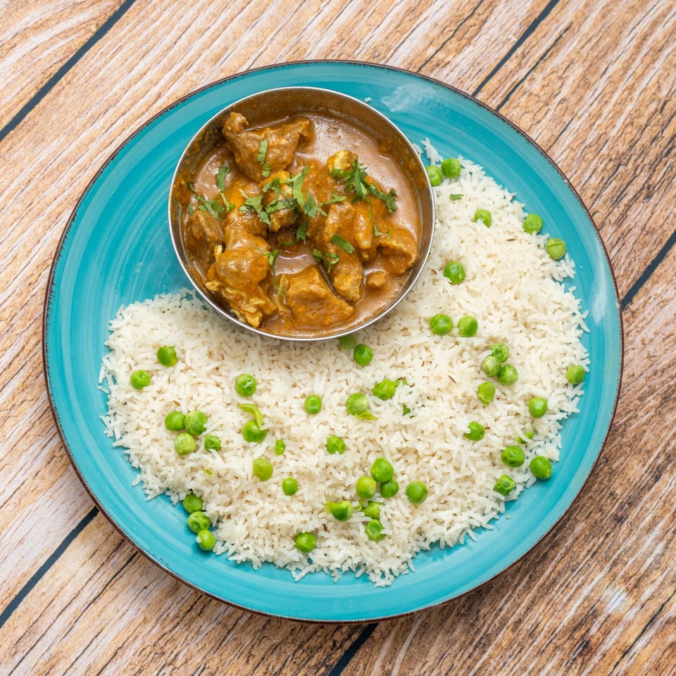 A teal plate with a bowl of lamb curry and a mound of white rice with green peas.