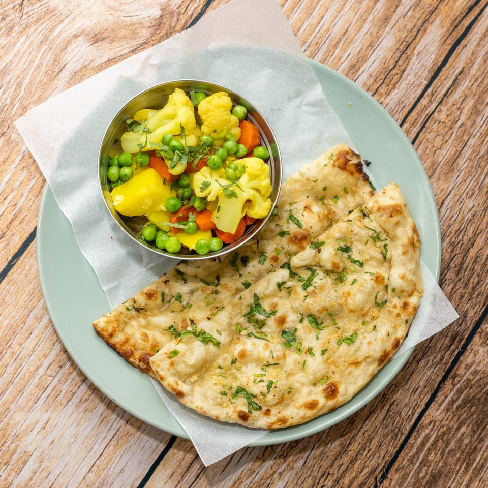 A light blue plate with garlic naan and a metal bowl of mixed vegetables on a wooden table.