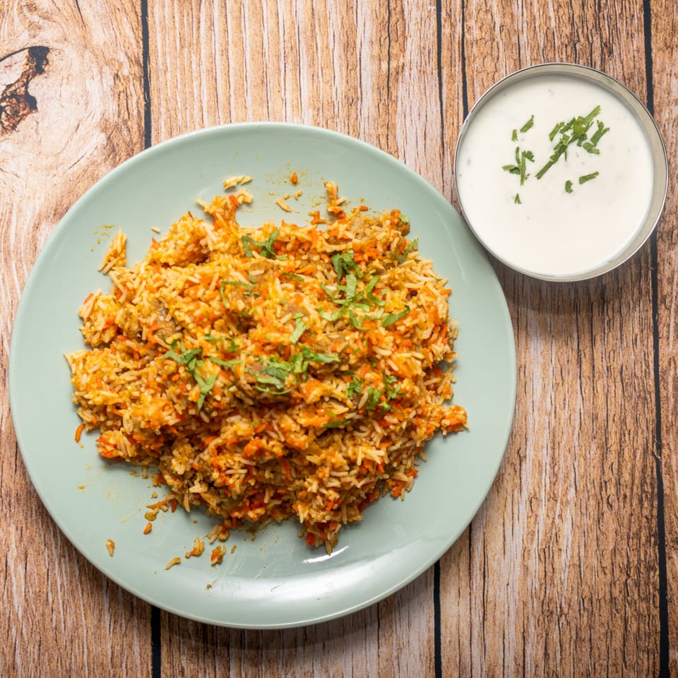 A plate of biryani with rice, carrots, and herbs, alongside a bowl of raita.