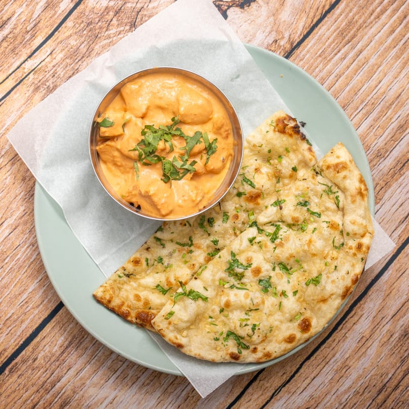 Butter chicken in a metal bowl with two pieces of naan bread on a light blue plate.